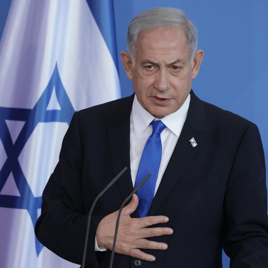 Benjamin Netanyahu wearing suit stands in front of flag of Israel