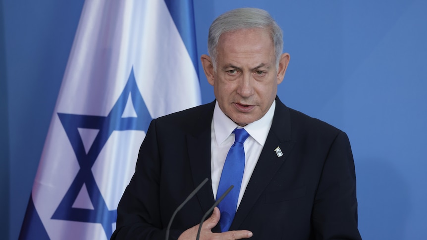 Benjamin Netanyahu wearing suit stands in front of flag of Israel
