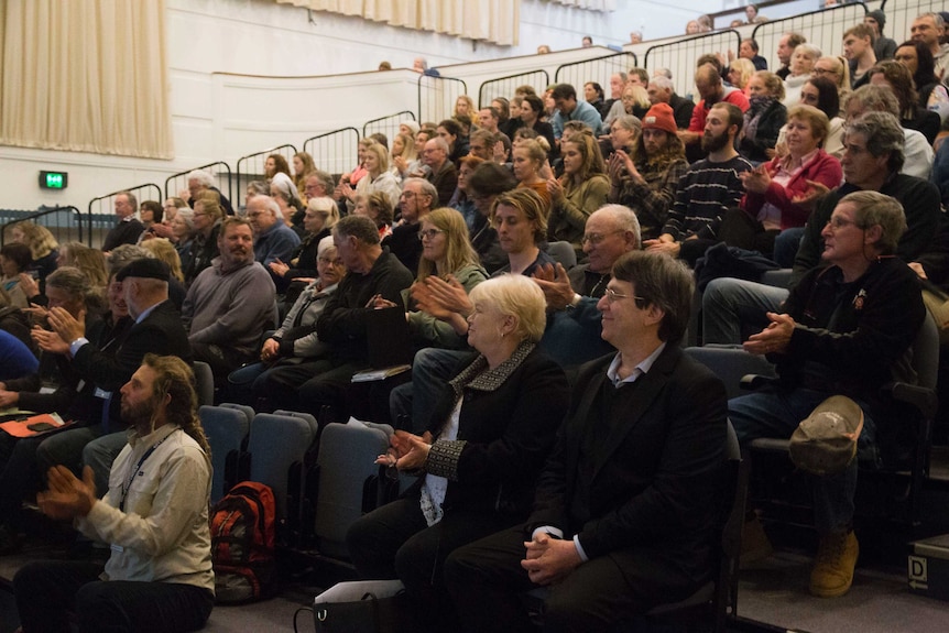 A mostly white crowd of mixed ages sits and claps on tiered seating inside a large theatre hall.