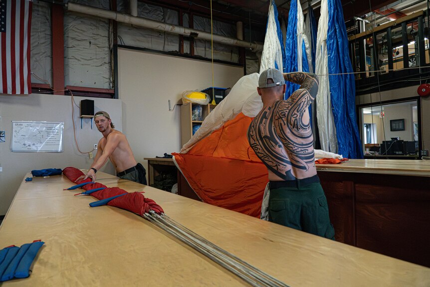 Two men work with parachutes along wooden work benches. One's back is heavily tattooed.