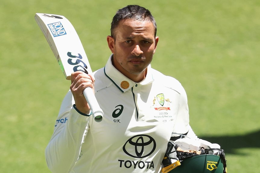 Usman Khawaja walks off with his helmet under one arm and holds up his bat