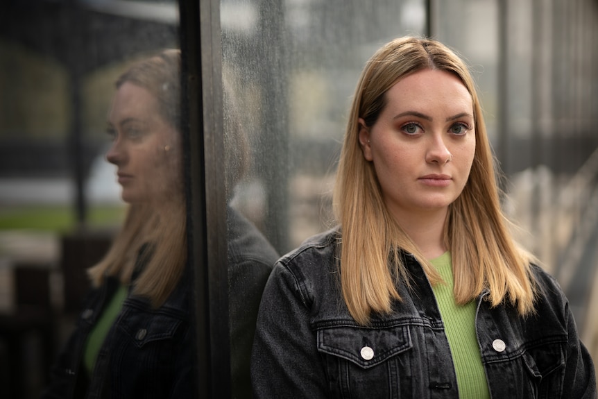 A woman leans against a window, showing her reflection. She stares at the camera with a neutral expression.