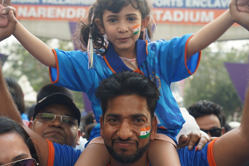 A young girl wearing an Indian cricket jersey and with the Indian flag painted on her face sits on a man's shoulders
