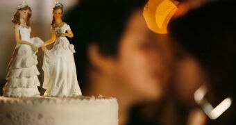 A wedding cake has two bride ornaments standing on top of it and a blurred same-sex couple kisses in the background.