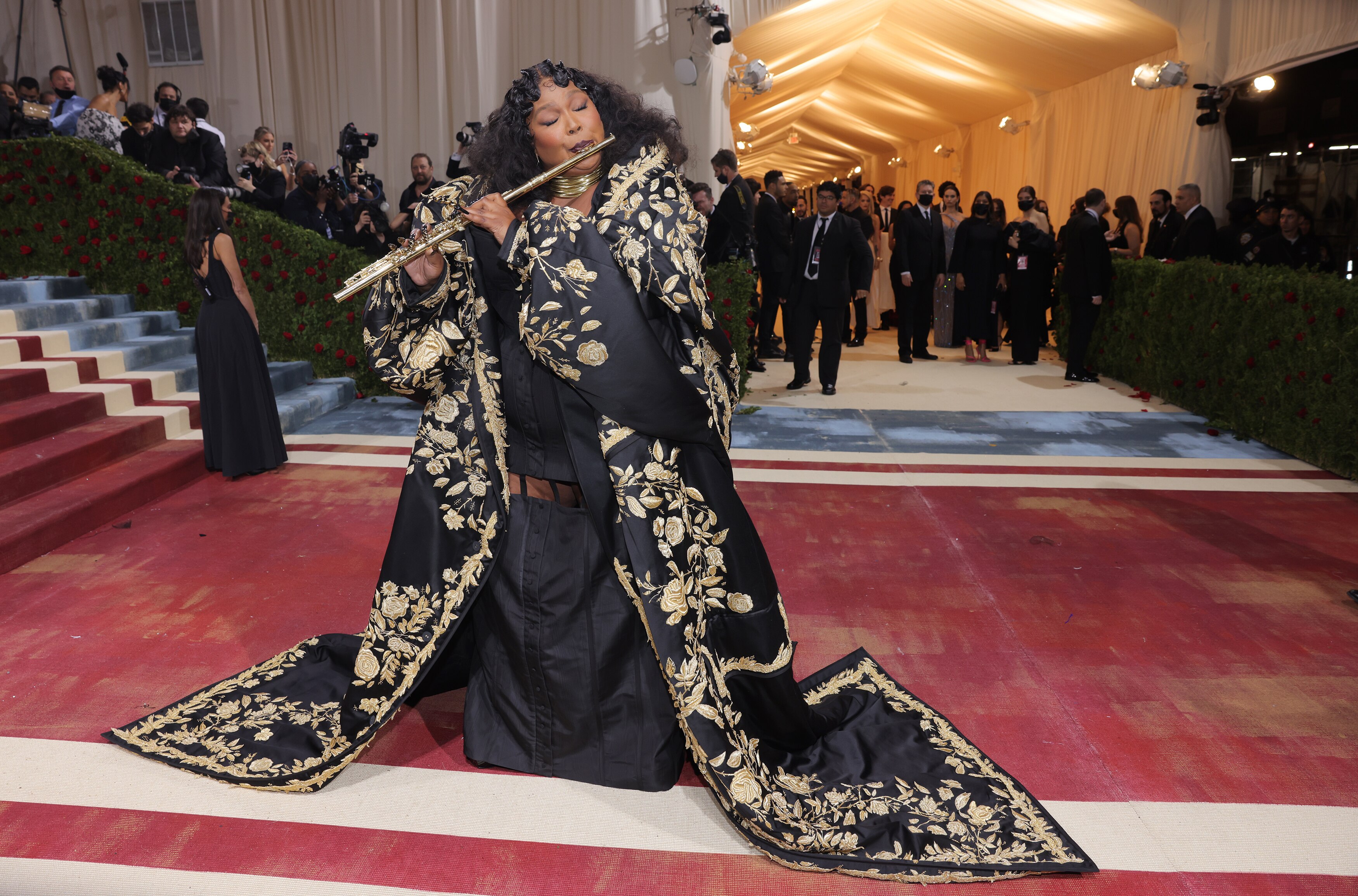 Lizzo, wearing a black and gold robe, plays a flute on the red carpet.