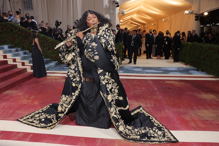Lizzo, wearing a black and gold robe, plays a flute on the red carpet.