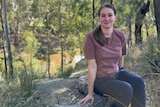 A woman in a purple tshirt and dark blue jeans sits on a fallen tree stump by a river in bushland