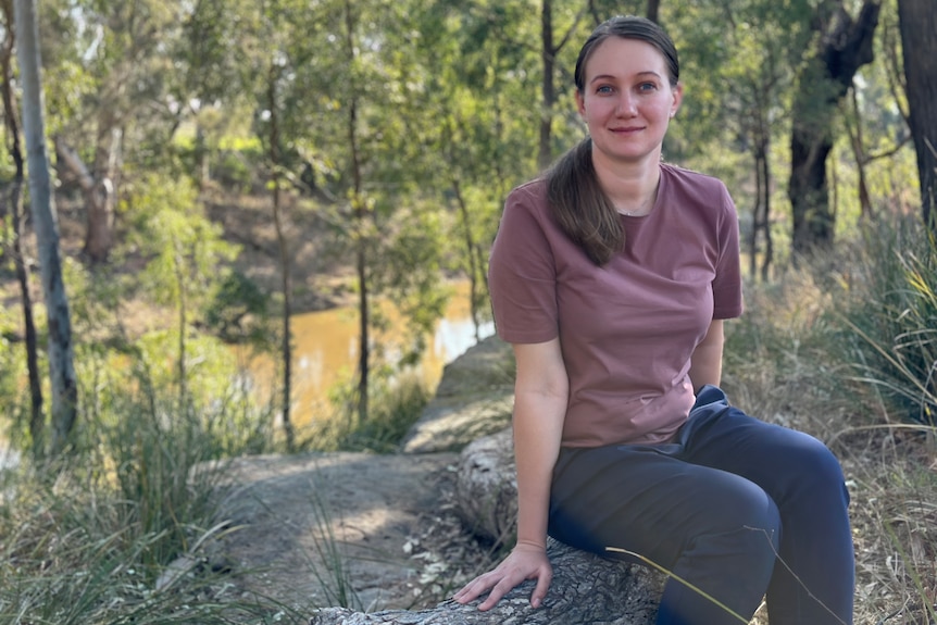 A woman in a purple tshirt and dark blue jeans sits on a fallen tree stump by a river in bushland