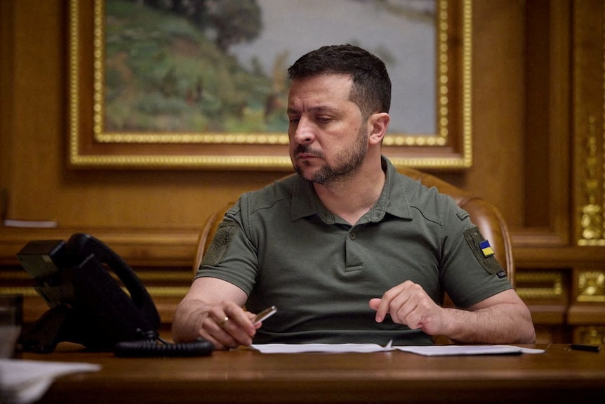 Volodymyr Zelenskyy, wearing a khaki polo shirt, sits at a desk looking at the desk phone