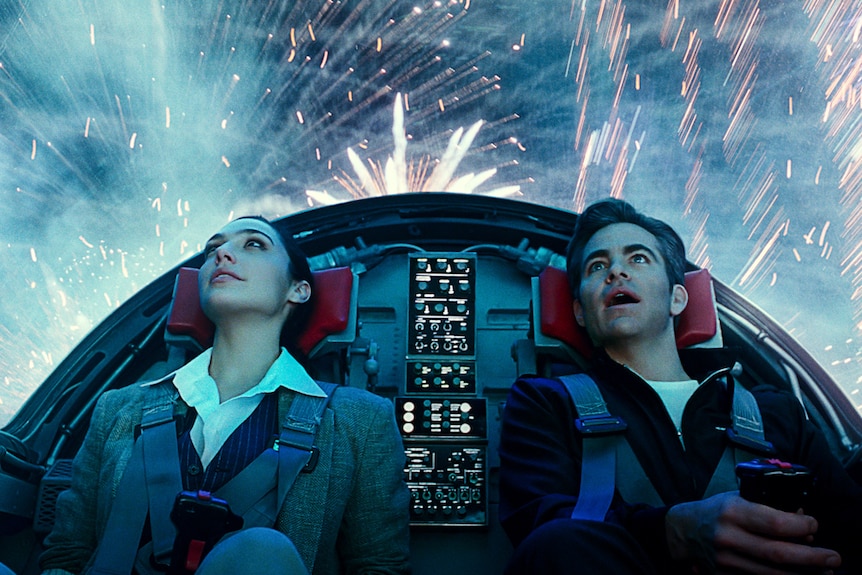 Gal Gadot and Chris Pine sit inside cockpit of spaceship with bright lights and fireworks outside around them and behind.