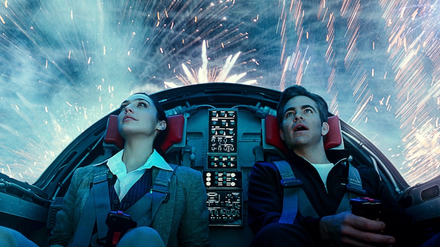 Gal Gadot and Chris Pine sit inside cockpit of spaceship with bright lights and fireworks outside around them and behind.