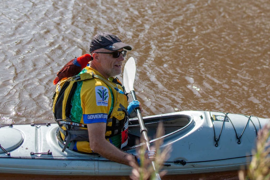 Older man in a kayak with a red parrot on his shoulder. 