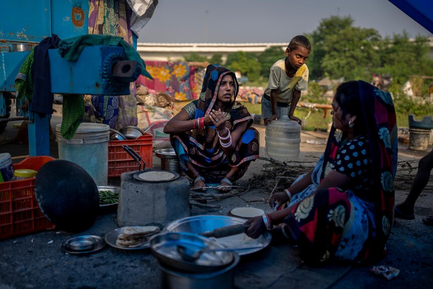 Two women sit on the road making flatbread with their hands as a child watches on. 