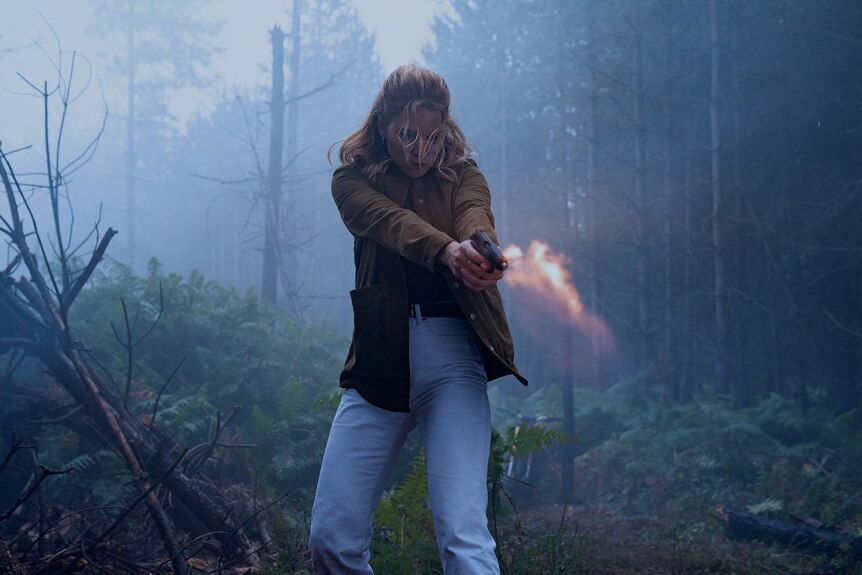 Blonde woman wearing a brown jacket and pale blue jeans shoots a handgun in a misty forest.