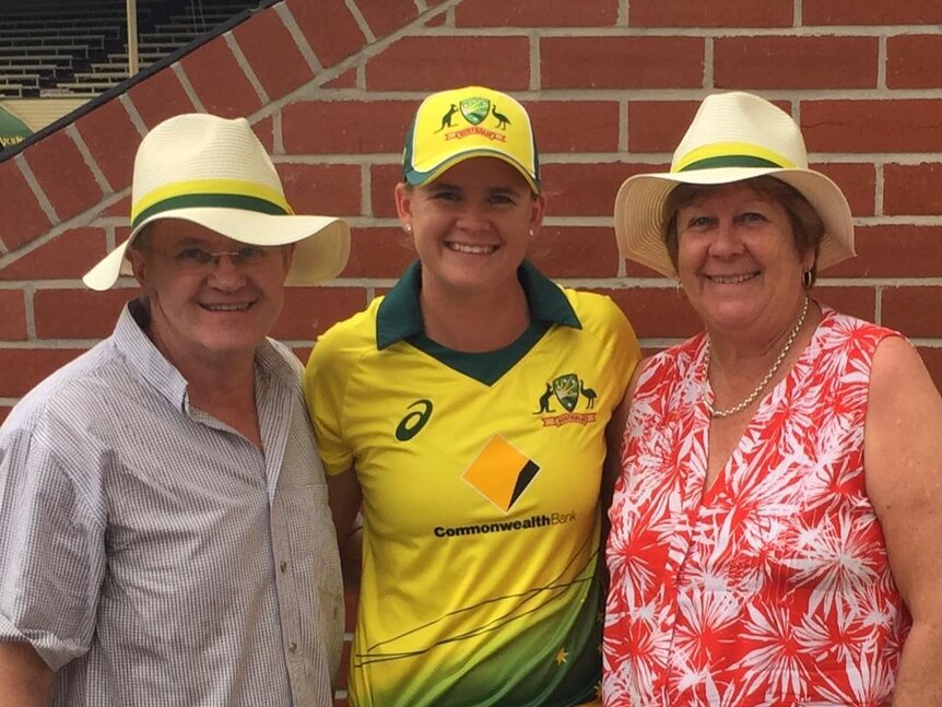 Jess Jonassen stands in her Australian kit, while her parents stand either side of her smiling.