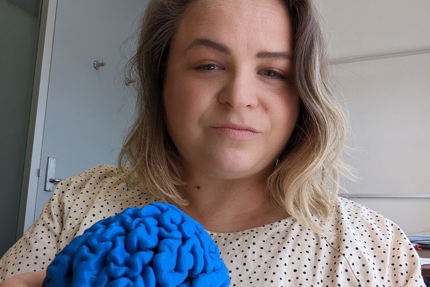 Sarah Hellewell, who has short blonde hair and is wearing a spotted cream top, poses for a photo holding a blue model of a brain