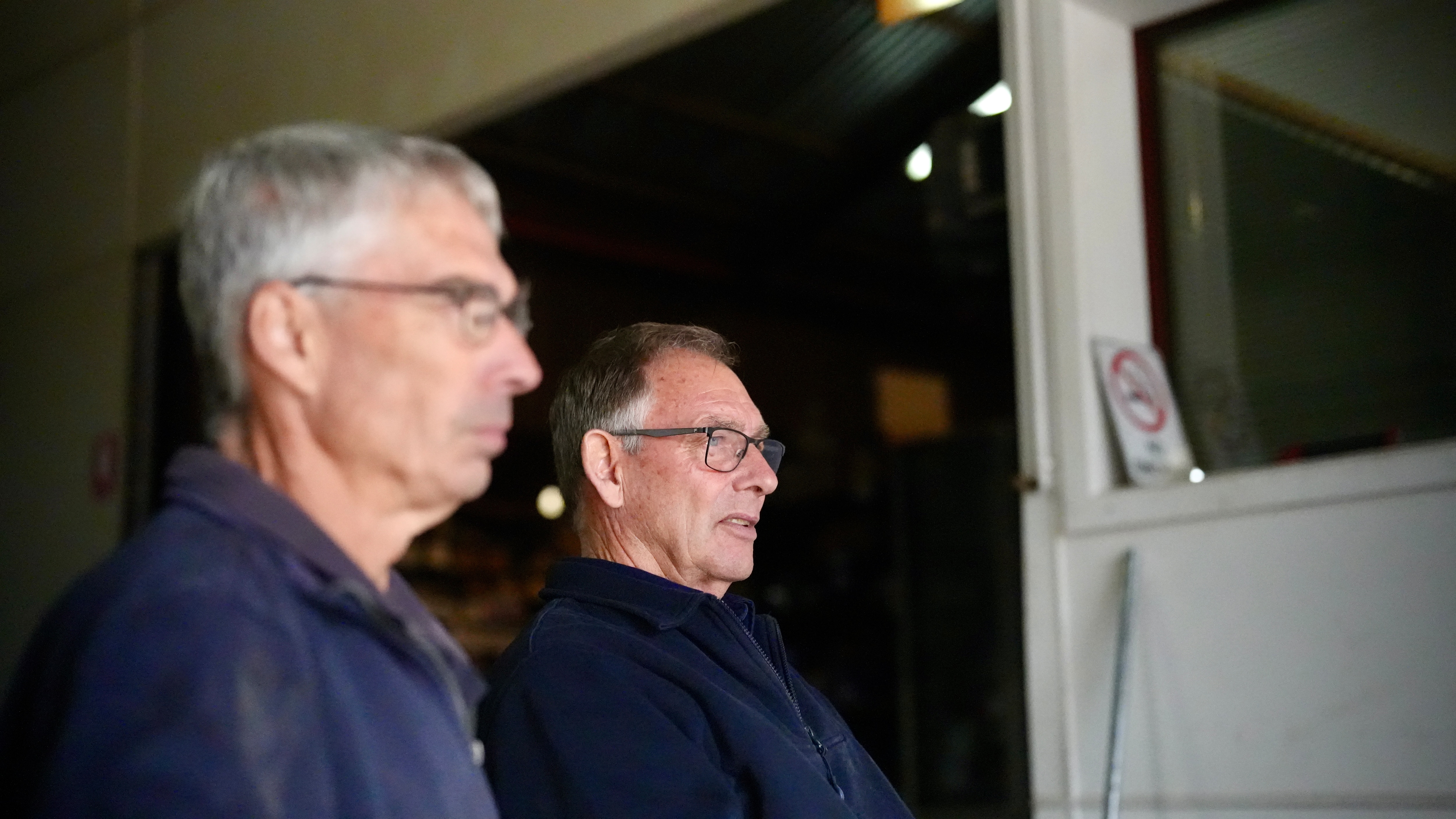 Dos hombres de mediana edad, uno enfocado y otro desenfocado, miran el taller mecánico a la derecha de la cámara.