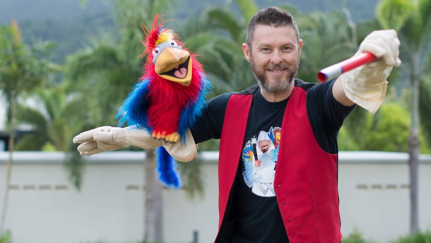A man in a red vest stands with a magician's wand in one hand and a bird puppet in the other.