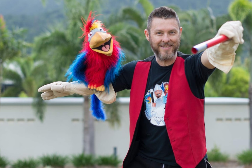 A man in a red vest stands with a magician's wand in one hand and a bird puppet in the other.