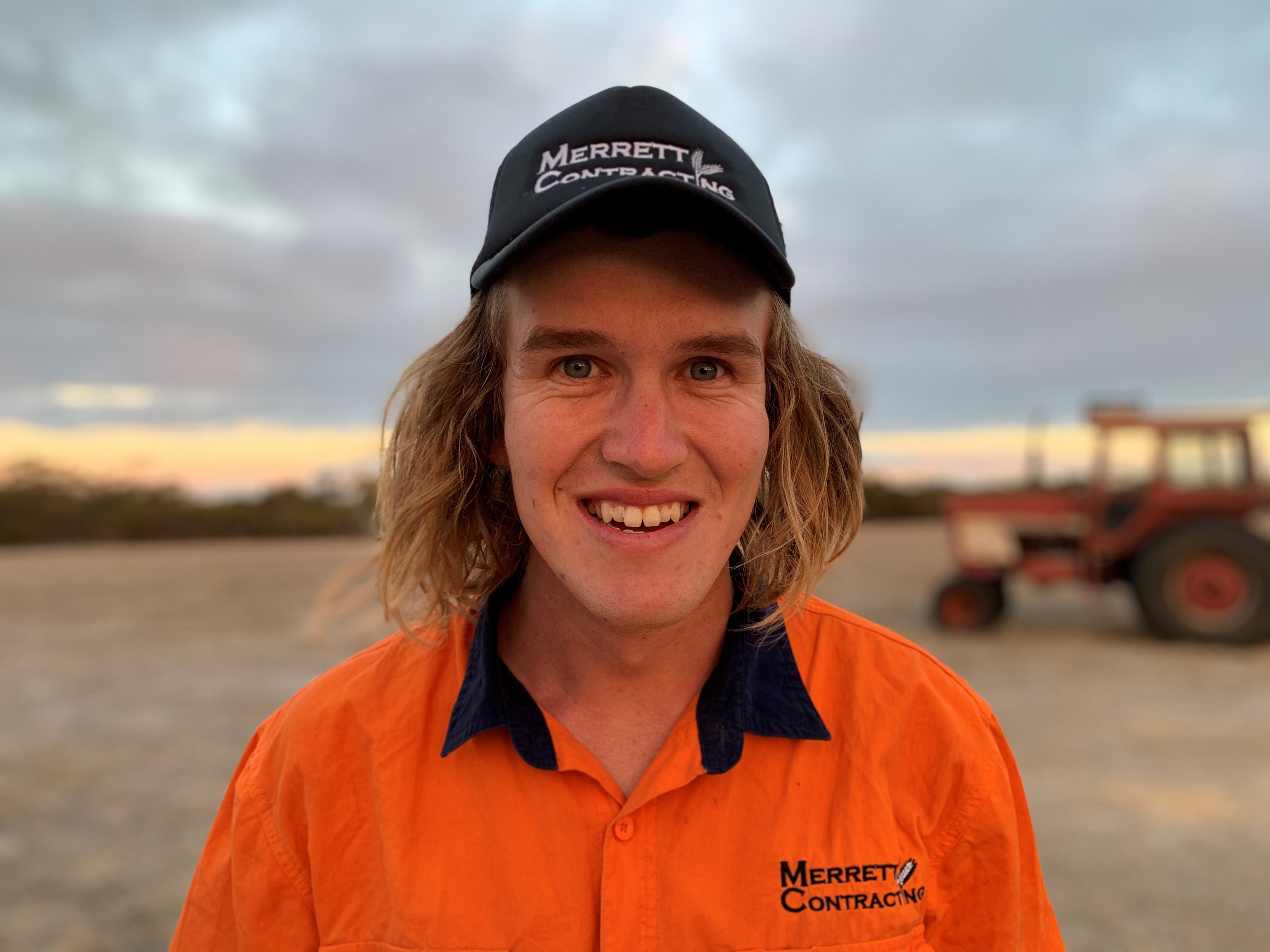 A white man with shoulder-length blonde hair looks to the camera and smiles. He wears an orange high-vis top and black cap.