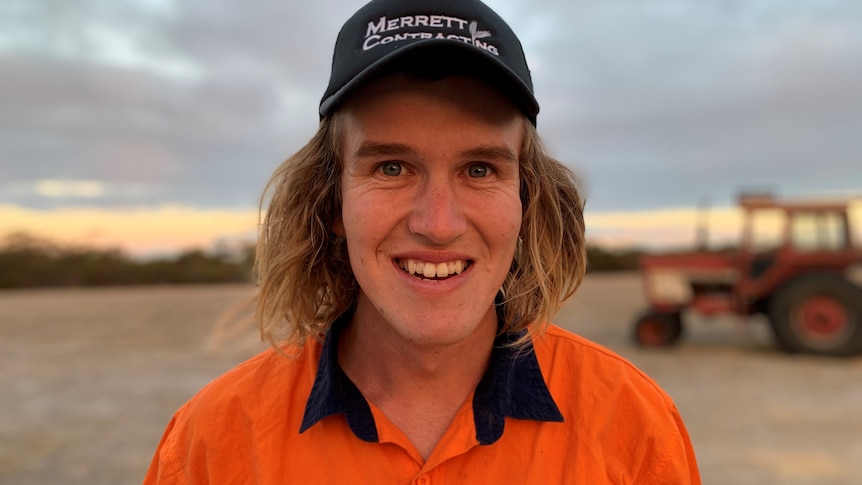 A white man with shoulder-length blonde hair looks to the camera and smiles. He wears an orange high-vis top and black cap.