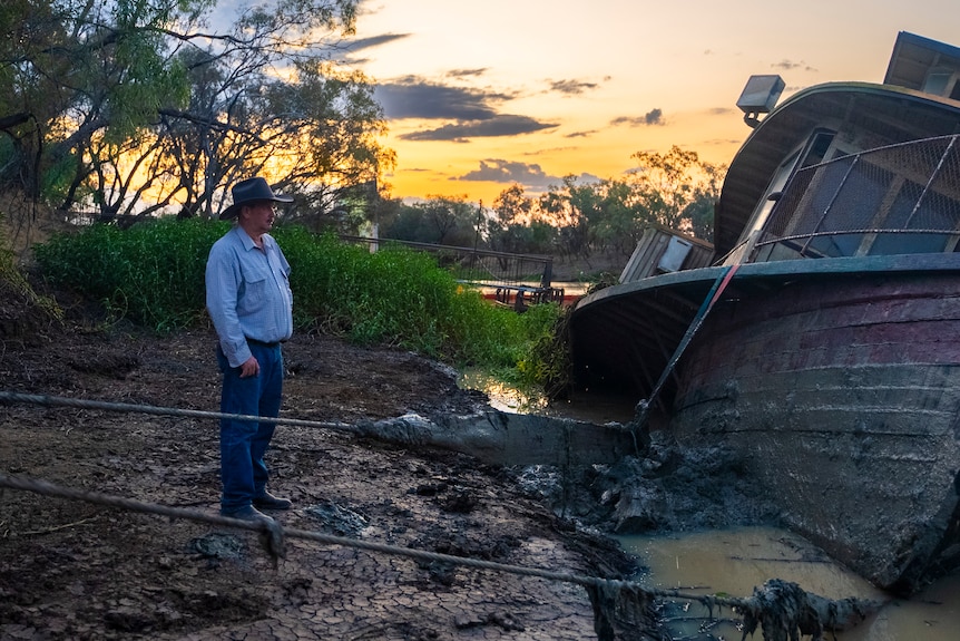 A man in a blue shirt and cowboy hat looks solemnly upon a half sunken boat buried by mud and brown water.
