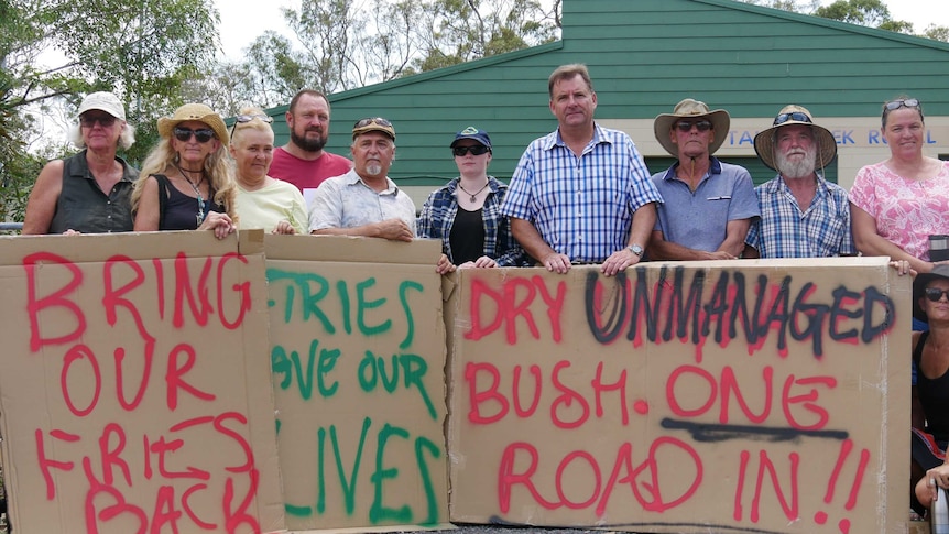 Ten people stand in a line in front of the Captain Creek Rural Fire station holding signs of protest 'Bring our firies back'