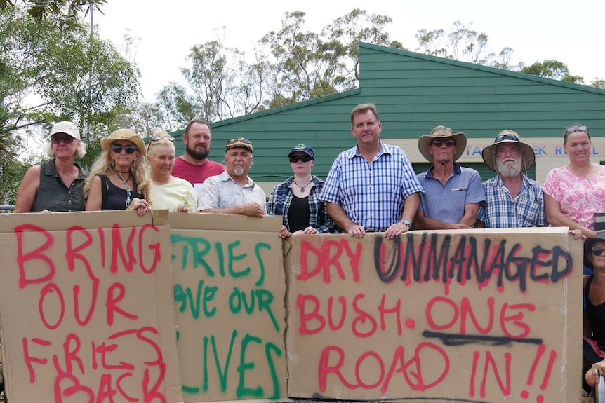 Ten people stand in a line in front of the Captain Creek Rural Fire station holding signs of protest 'Bring our firies back'