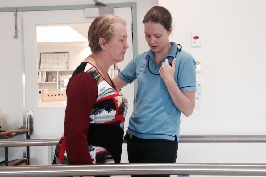 A middle-aged woman walks between two support bars, helped by a nurse, while recovering in hospital.