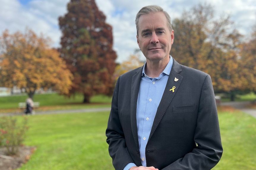 Michael Ferguson wearing a grey suit and blue shirt stands in a park with autumn trees in the background