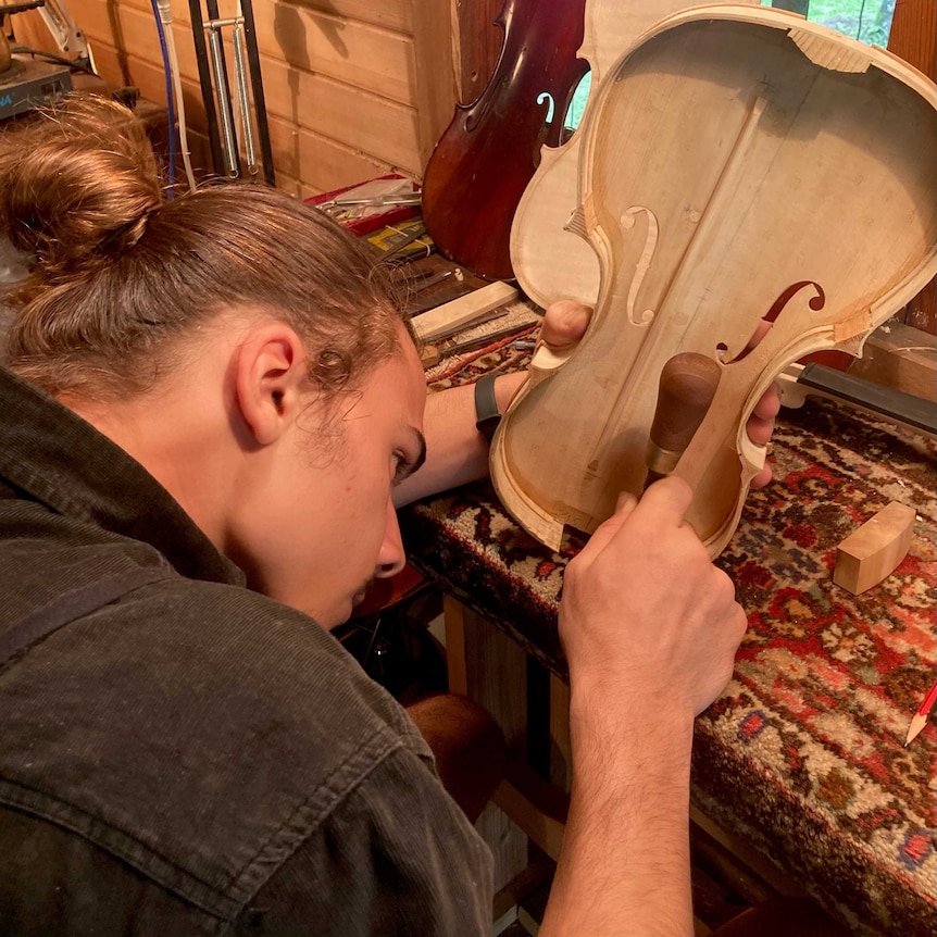 a man focuses intently on his work bench, holding a chisel and beginnings of a violin