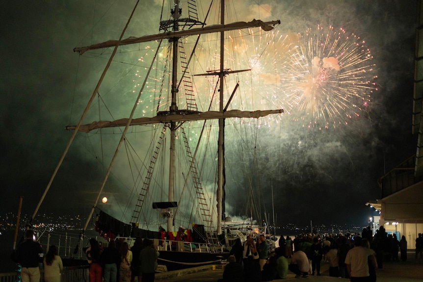 A wide shot of people outside looking up at fireworks in the night sky, beyond where a ship sits docked.