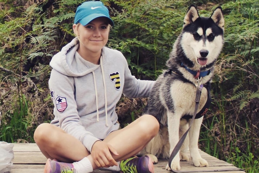 A young woman in sports clothes and a blue cap sits on a timber bench beside a husky, smiling.