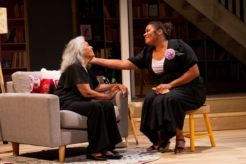 An older woman with grey hair sits on a couch; a younger woman sits near her and touches her face tenderly, smiling.