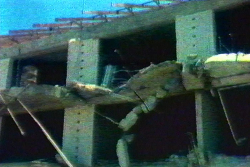 Blurry TV still, building exterior falling to pieces, concrete and steel hanging in the air, blue sky behind.