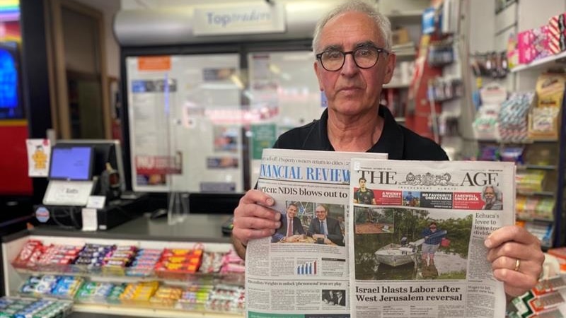 A man with grey hair and glasses holds up two newspapers in a shop