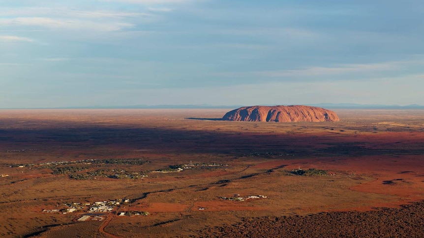 A picture of Uluru from the air, with blue skies and long stretches of red earth.