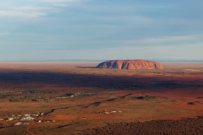 A picture of Uluru from the air, with blue skies and long stretches of red earth.