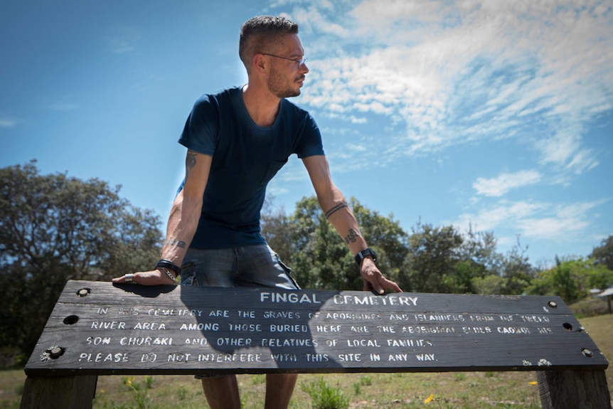 ABC RN Presenter for Awaye! Daniel Browning is leaning on a signpost of a cemetery in his hometown, Fingal