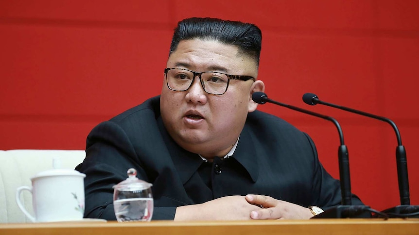 North Korean leader Kim Jong Un sitting at a desk with microphones in front of him and a red background behind him