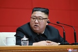 North Korean leader Kim Jong Un sitting at a desk with microphones in front of him and a red background behind him