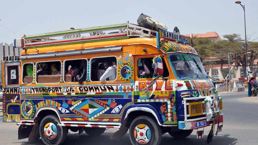 Small bus painted with bright colours on a yellow background. The bus is packed with people and sitting in the street.