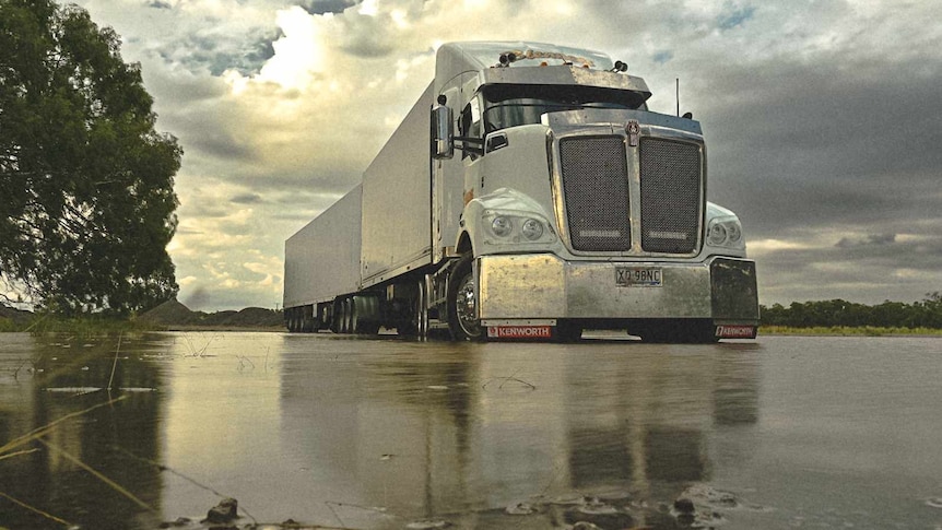 A grey truck drives along a wet, isolated-looking road.