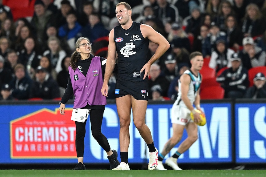 A Carlton AFL player grimaces as he is assisted by a trainer after sustaining an injury.