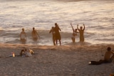 People in the water and on the sand at a crowded beach during sunset.