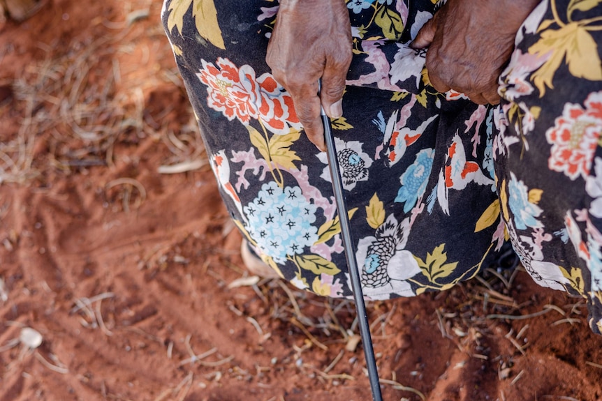 A woman's hands rest on her patterned print skirt, as she draws in red sand with a stick.