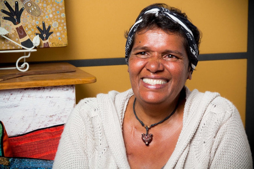 Aunty Denise Bloomfield sits in front of a yellow wall smiling.