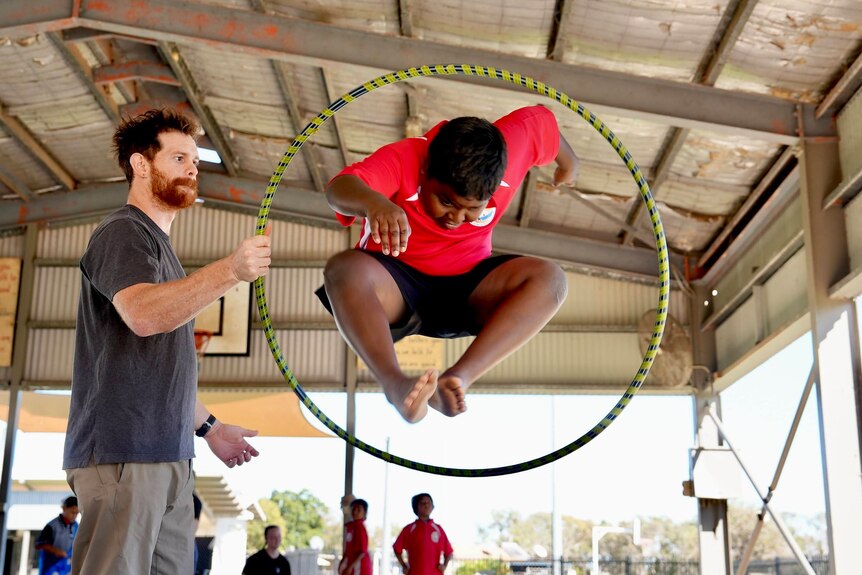 A young school student, wearing a red shirt, jumps through a yellow and blue hula-hoop, being held by a male circus trainer. 