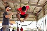 A young school student, wearing a red shirt, jumps through a yellow and blue hula-hoop, being held by a male circus trainer. 