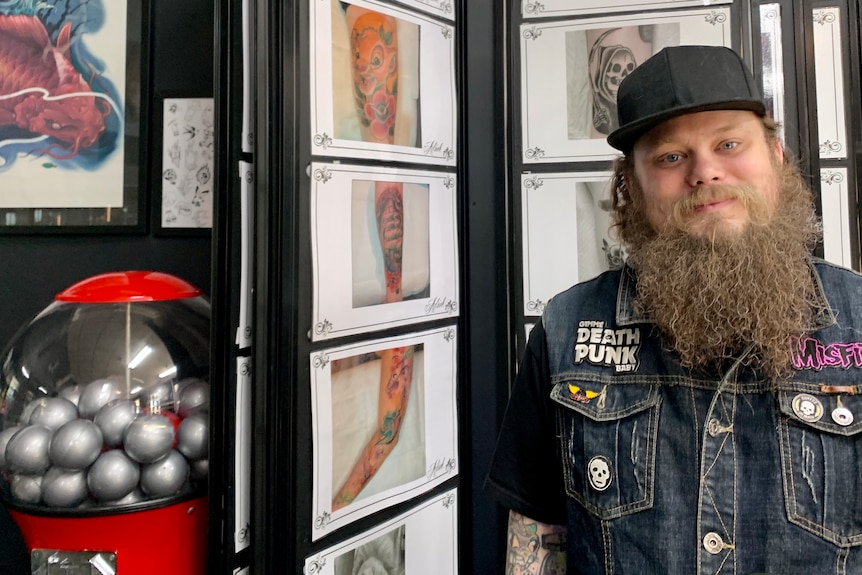 A man with a beard stands in front of a wall of tattoo designs.